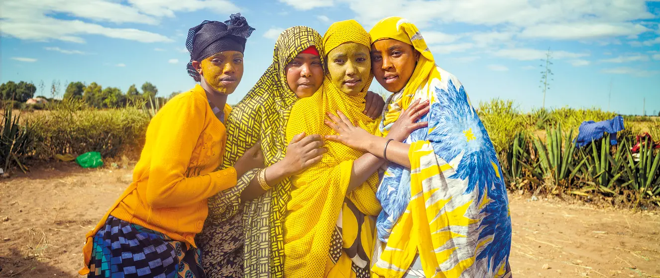 Vier Mädchen mit gelb gefärbten Gesichtern und gelb-blauen Tüchern stehen vor einem Feld und umarmen sich