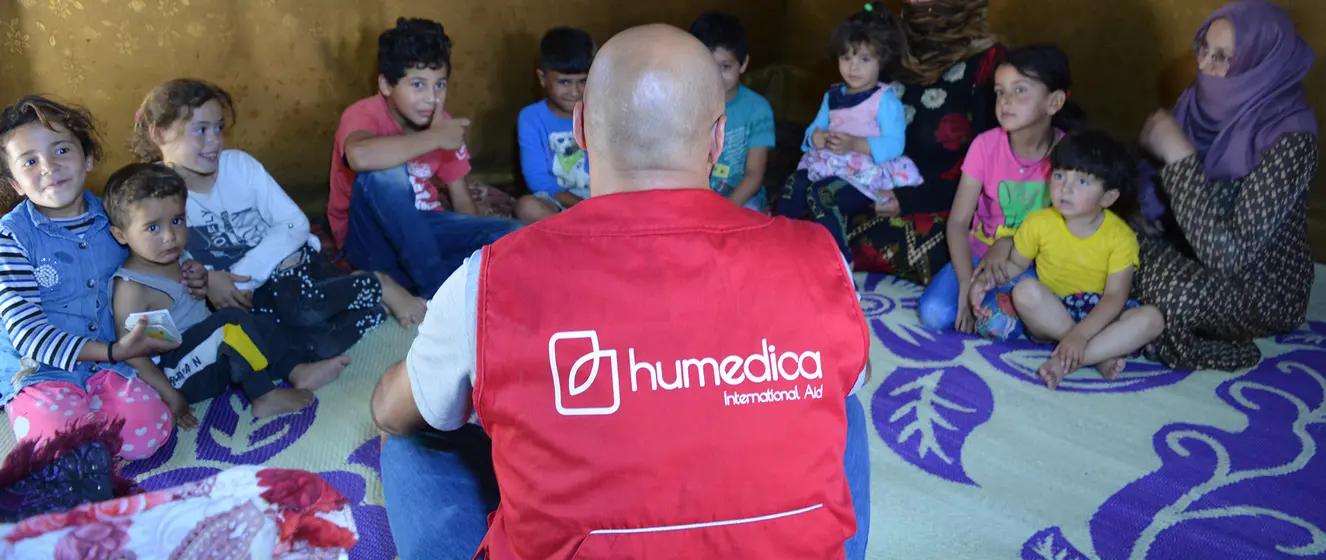 Kinder sitzen im Kreis und schauen zu einem Mann, der mit roter humedica-Weste vor ihnen sitzt