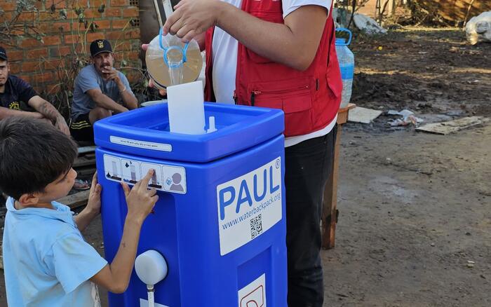 Ein Junge steht vor einem blauen rechteckigem Wasserfilter in den ein Mann mit roter humedica-Weste verschmutztes Wasser einfüllt