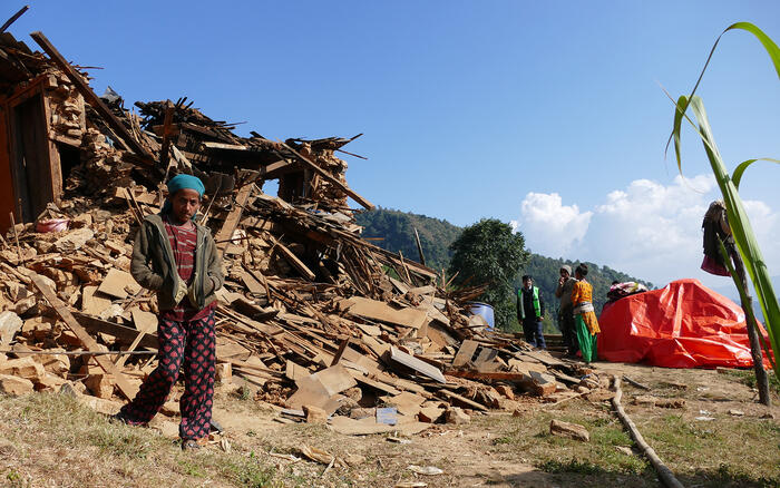 Ein Mädchen steht vor einem zerstörten Haus nach einem Erdbeben in Nepal, rechts davon stehen Menschen neben einer orangefarbenen Plane