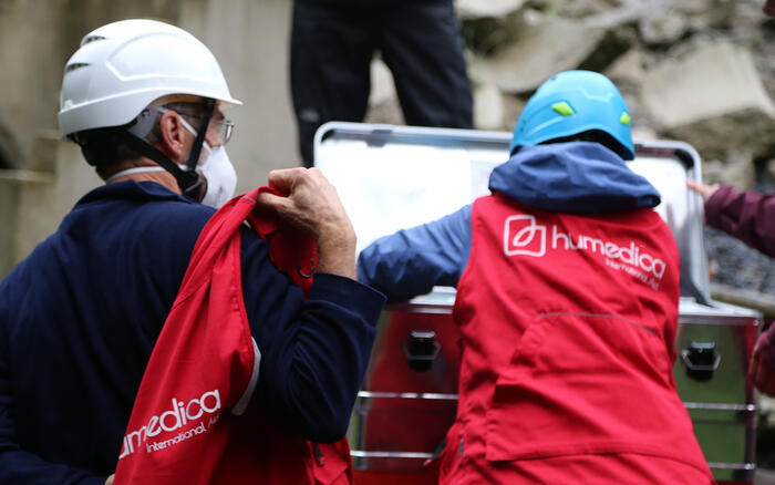 Zwei Menschen stehen mit roten humedica Westen vor einer grauen Metallkiste, einer greift hinein