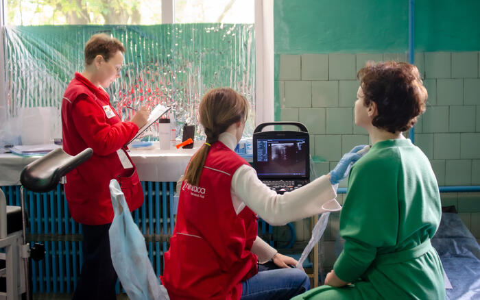 Eine Ärztin mit roter humedica Weste untersucht mit einem Ultraschall-Gerät den Hals einer Frau, die mit dem Rücken zur Kamera sitzt. Eine weitere Frau steht daneben und macht Notizen.