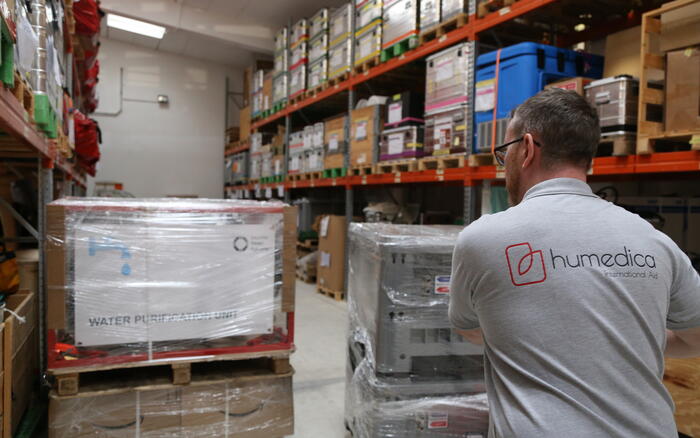 Ein Mann mit einem grauen humedica T-Shirt schiebt auf einer Sackkarre eine Palette verpackter Kisten durch ein Lager