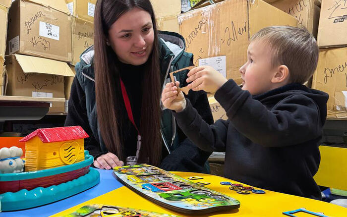 A woman sits smiling next to a small child who is showing her something, in front of them is a puzzle on the table, in the background are stacked cardboard boxes