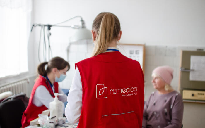 In einem Behandlungszimmer sitzt eine Ärztin mit einer Patientin am Tisch, eine Frau mit humedica-Weste steht mit dem Rücken zur Kamera im Vordergrund