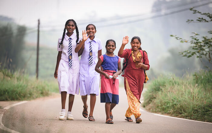 Vier Mädchen, zwei davon in weißer Schuluniform, laufen eine Straße in Sri Lanka entlang und winken