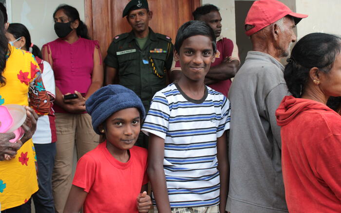 Ein Junge und ein Mädchen stehen in einer Menschenmenge vor einem Hilfezentrum in Sri Lanka und lächeln in die Kamera