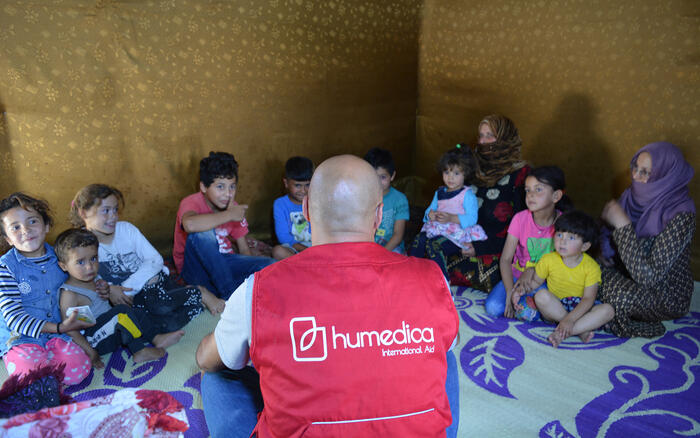 Kinder sitzen im Kreis und schauen zu einem Mann, der mit roter humedica-Weste vor ihnen sitzt