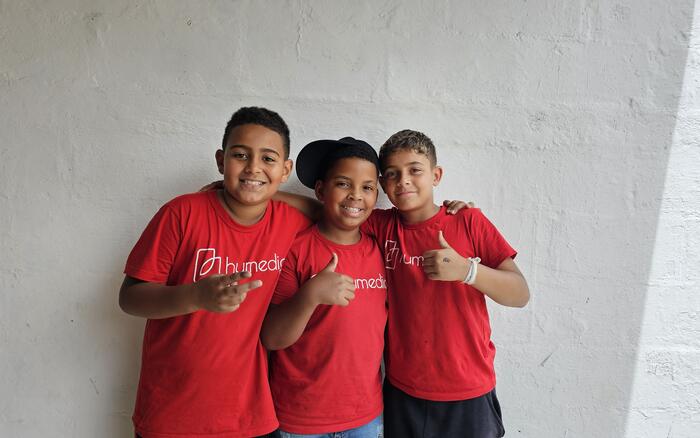 Drei brasilianische Jungs stehen mit roten humedica-T-Shirts vor einer weißen Wand und halten die Daumen nach oben.