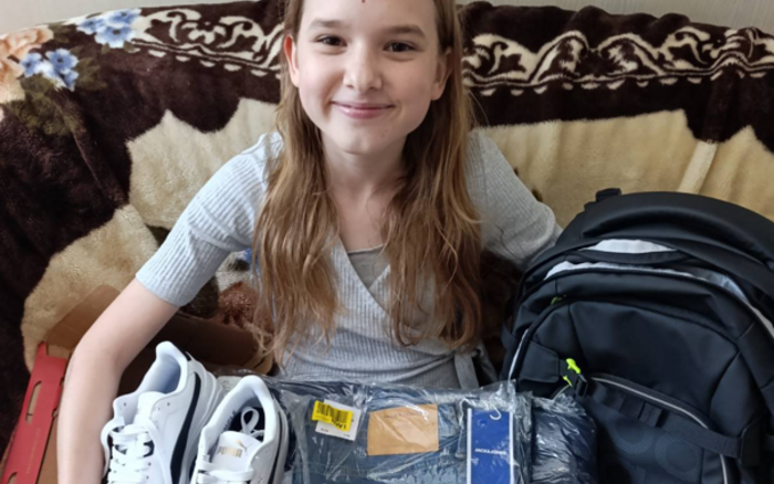 Ein Mädchen sitzt lächelnd auf einem Sofa und hält Schuhe, Kleidung und einen Rucksack auf dem Schoß