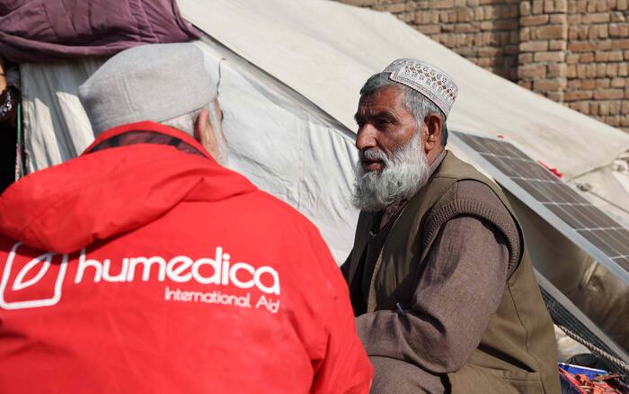 Ein Mann in einer roten humedica Jacke schaut einen anderen afghanischen Mann an. Sie sitzen sich gegenüber und reden.