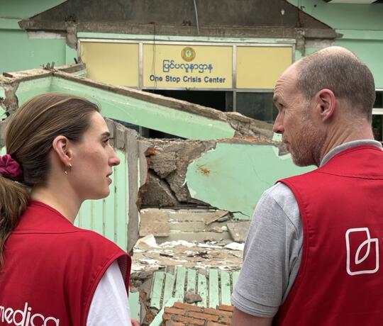 Eine Frau und ein Mann mit roten humedica-Westen stehen vor einem zerstörten Haus