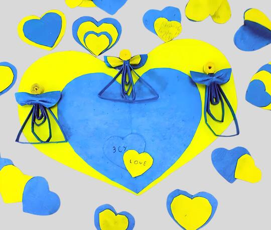 Unterschiedlich große blaue und gelbe Papierherzen liegen neben- und übereinander auf einem Tisch, drei aus Papier gefaltete Engel darüber