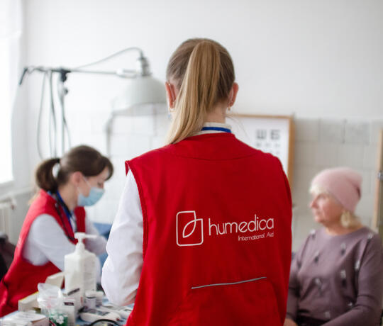 In einem Behandlungszimmer sitzt eine Ärztin mit einer Patientin am Tisch, eine Frau mit humedica-Weste steht mit dem Rücken zur Kamera im Vordergrund