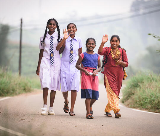 Vier Mädchen, zwei davon in weißer Schuluniform, laufen eine Straße in Sri Lanka entlang und winken