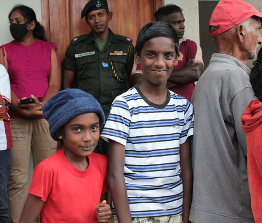 Ein Junge und ein Mädchen stehen in einer Menschenmenge vor einem Hilfezentrum in Sri Lanka und lächeln in die Kamera
