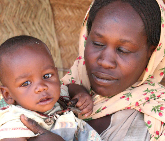Porträt einer Mutter, die ihr Baby, welches in die Kamera schaut, auf dem Arm hält.