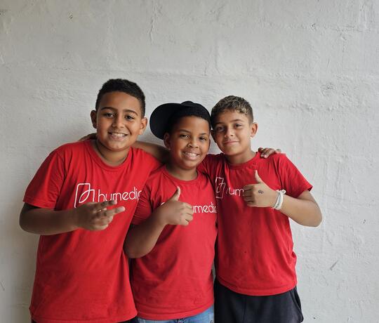 Drei brasilianische Jungs stehen mit roten humedica-T-Shirts vor einer weißen Wand und halten die Daumen nach oben.