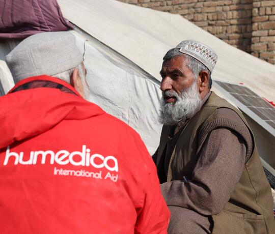 Ein Mann in einer roten humedica Jacke schaut einen anderen afghanischen Mann an. Sie sitzen sich gegenüber und reden.