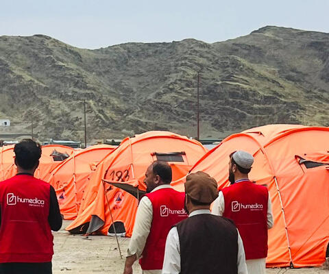 Drei Männer mit roten humedica-Westen gehen an orangefarbenen Zelten in einem Geflüchtetencamp in Afghanistan vorbei