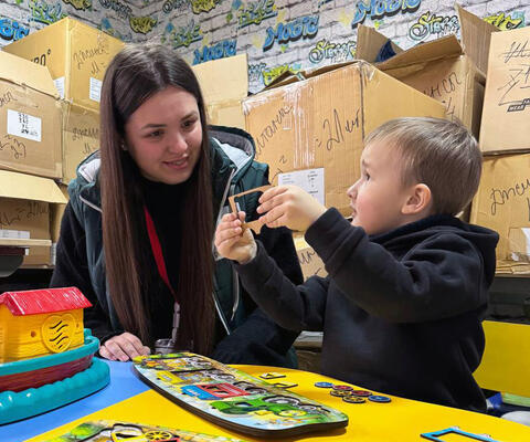 Eine Frau sitzt lächelnd neben einem kleinen Kind, das ihr etwas zeigt, vor ihnen liegt ein Puzzle auf dem Tisch, im Hintergrund sind gestapelte Kartons