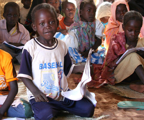 Im Vordergrund kniet ein Junge auf dem Boden mit einem Stift in der rechten und Papier in der linken Hand. Dabei schaut er in die Kamera. Um ihn herum sitzen noch weitere Kinder auf dem Boden.