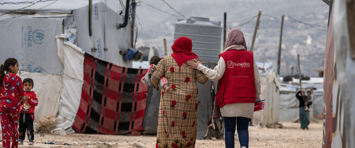 Frau mit roter humedica-Weste laeuft neben Frau, die ein Kind auf dem Arm traegt, durch ein Gefluechtetenlager im Libanon.