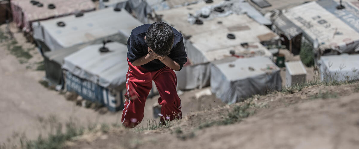 Ein Junge steht gebückt mit den Händen vor den Augen vor einem Geluechtetencamp