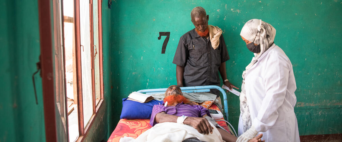 Ein Mann und eine Ärztin stehen an einem Krankhausbett, in dem eine Person liegt