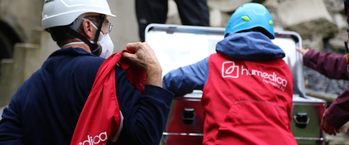 Zwei Menschen stehen mit roten humedica Westen vor einer grauen Metallkiste, einer greift hinein