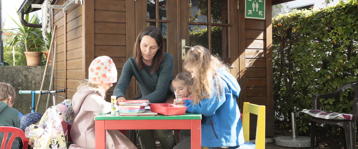 Eine Erzieherin sitzt mit drei Kindern an einem Tisch vor einem Gartenhäuschen