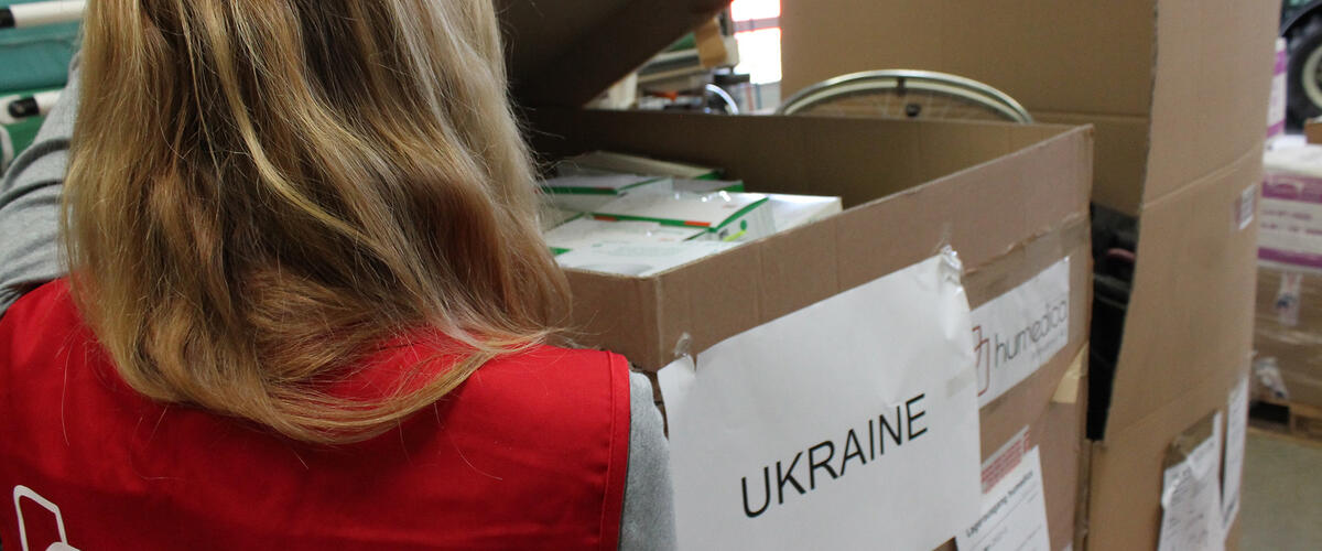 Frau mit roter humedica-Weste steht vor einem großen Karton mit der Aufschrift Ukraine, gefüllt mit kleineren Medikamentenpackungen