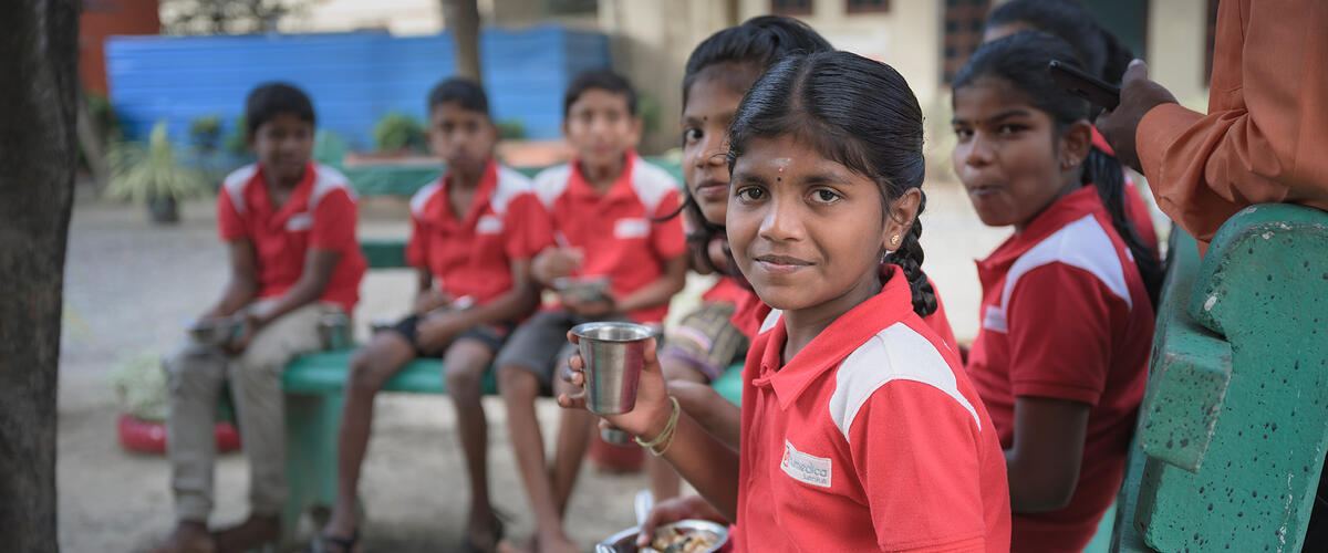 Ein Schulmädchen mit rotem Shirt sitzt mit ihren Mitschülern in einem Pausenhof in Sri Lanka auf einer grünen Bank, hält eine Metallschüssel in den Händen und schaut in die Kamera