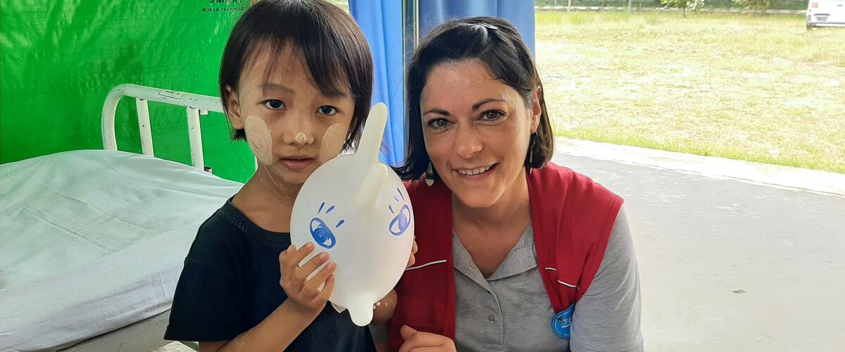 Ein Kind hält einen mit zwei Augen bemalten Luftballon (aufgeblasener Handschuh) in den Händen, daneben hockt eine medizinische Helferin mit roter humedica-Weste