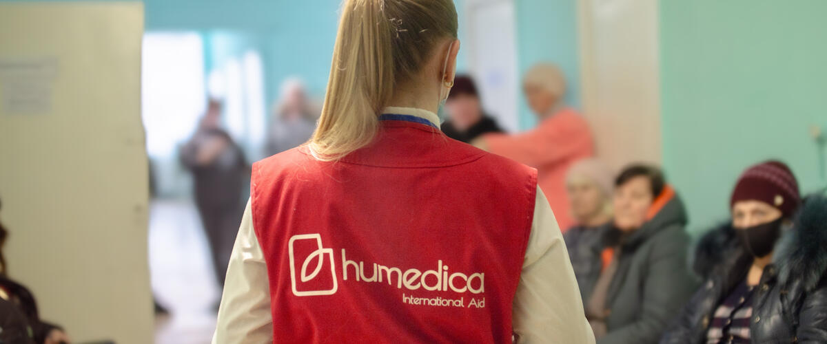 Eine Frau mit roter humedica Weste steht mit dem Rücken zur Kamera vor einer Gruppe Menschen in einer Art Wartezimmer.