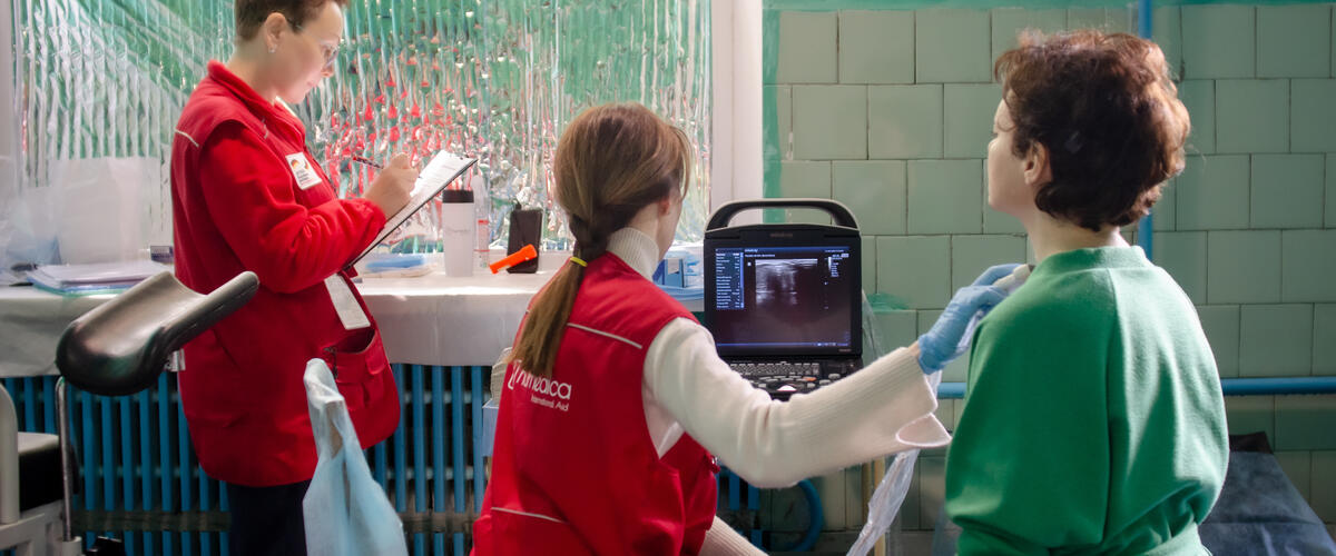 A doctor in a red humedica vest uses an ultrasound device to examine the neck of a woman sitting with her back to the camera. Another woman stands next to her and takes notes.