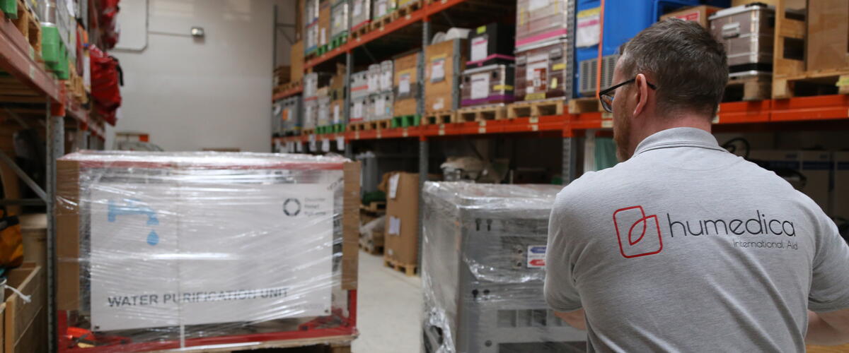 Ein Mann mit einem grauen humedica T-Shirt schiebt auf einer Sackkarre eine Palette verpackter Kisten durch ein Lager