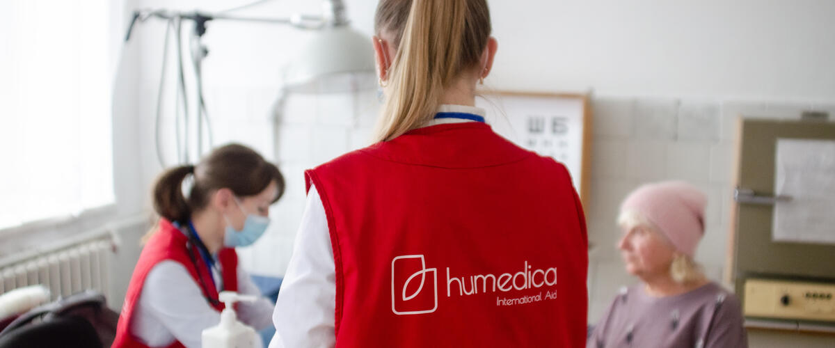 In einem Behandlungszimmer sitzt eine Ärztin mit einer Patientin am Tisch, eine Frau mit humedica-Weste steht mit dem Rücken zur Kamera im Vordergrund