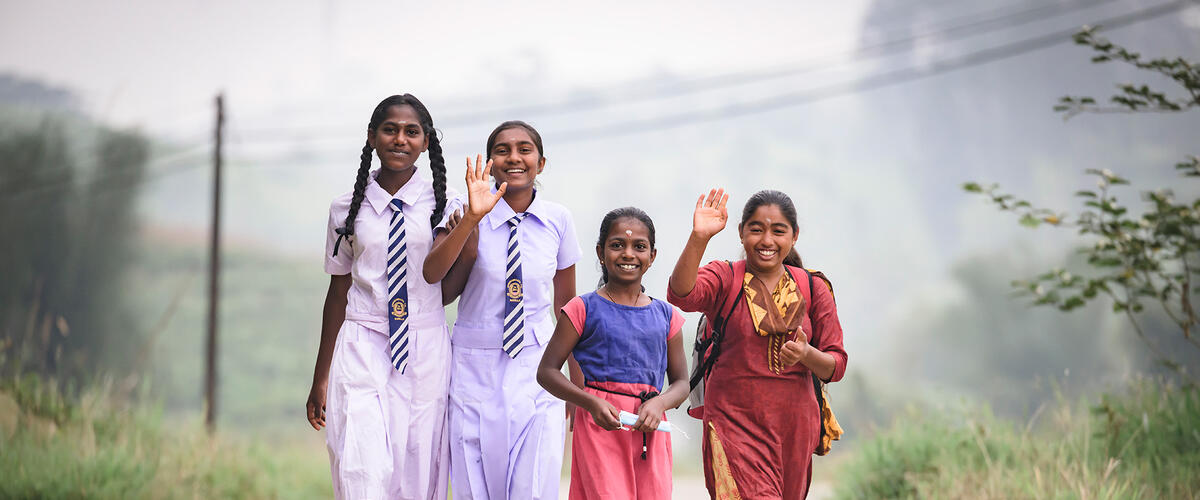 Vier Mädchen, zwei davon in weißer Schuluniform, laufen eine Straße in Sri Lanka entlang und winken