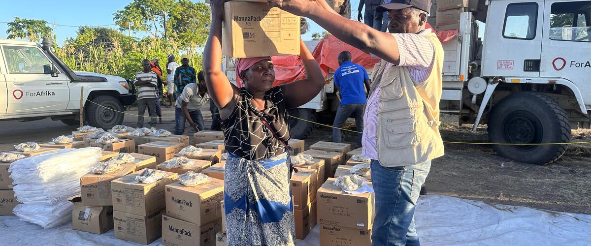 Eine Frau trägt zwei Kartons mit Lebensmitteln auf dem Kopf, ein Mann hilft beim Stabilisieren