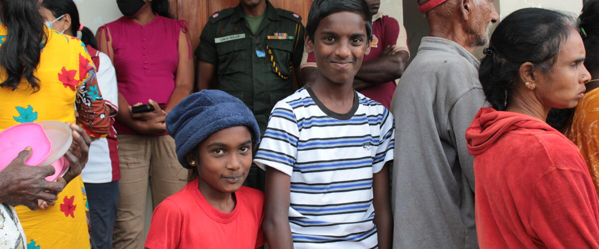 Ein Junge und ein Mädchen stehen in einer Menschenmenge vor einem Hilfezentrum in Sri Lanka und lächeln in die Kamera
