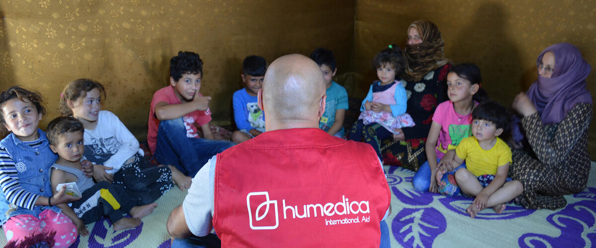 Kinder sitzen im Kreis und schauen zu einem Mann, der mit roter humedica-Weste vor ihnen sitzt