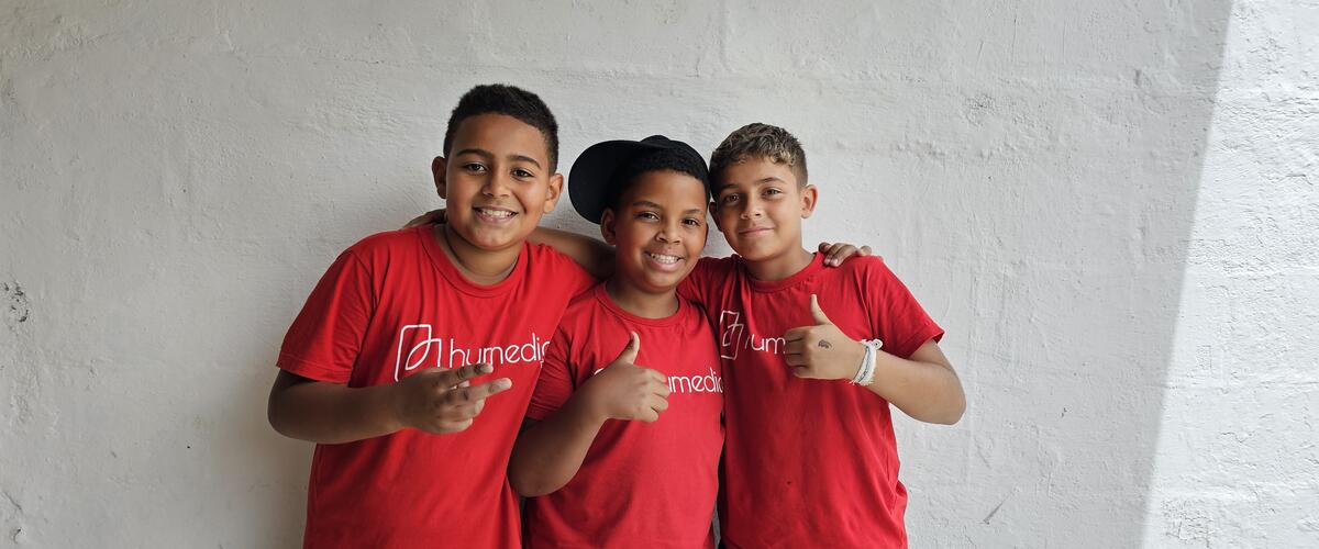 Drei brasilianische Jungs stehen mit roten humedica-T-Shirts vor einer weißen Wand und halten die Daumen nach oben.