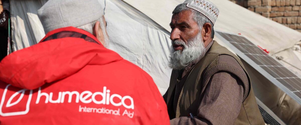 Ein Mann in einer roten humedica Jacke schaut einen anderen afghanischen Mann an. Sie sitzen sich gegenüber und reden.