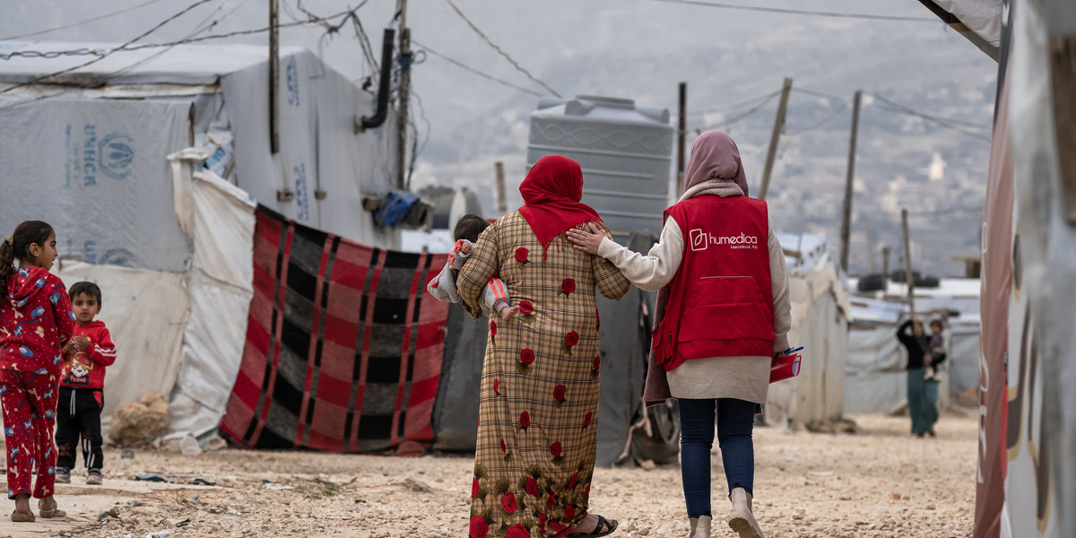 Frau mit roter humedica-Weste laeuft neben Frau, die ein Kind auf dem Arm traegt, durch ein Gefluechtetenlager im Libanon.