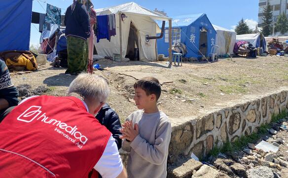humedica Einsatzkraft unterhält sich mit zwei Jungen, im Hintergrund ist das Zeltlager zu sehen, das als Notunterkunft nach dem Erdbeben dient.