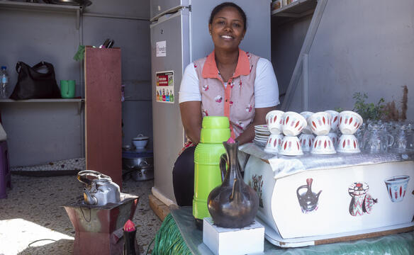 Eine Frau sitzt in einer kleinen Küche hinter Kaffee- und Teekannen und Tassen
