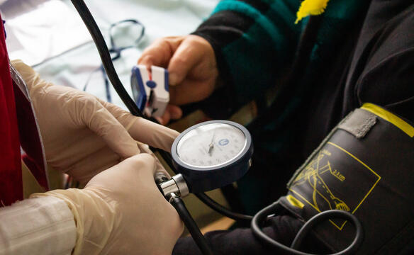 Zwei Hände mit Handschuhen halten ein Blutdruckmessgerät, welches an einem Arm befestigt ist. Die andere Hand der Person hat ein Sauerstoffmessgerät am Zeigefinger.