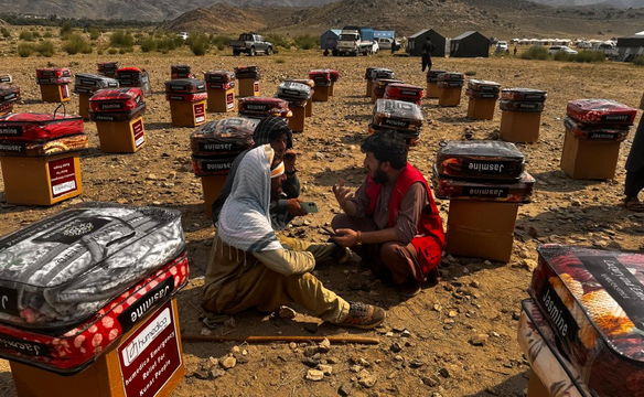 Drei Männer, davon einer mit roter humedica-Weste, sitzen inmitten von gestapelten Kisten und Decken, im Hintergrund Zelte und ein Gebirgszug