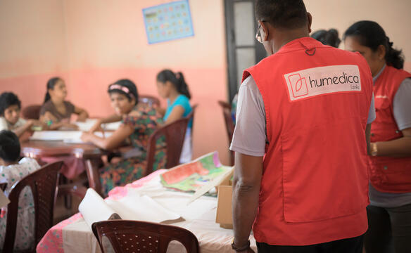 Zwei Menschen mit roter humedica Weste stehen an einem Tisch, im Hintergrund sind verschwommen Kinder zu sehen